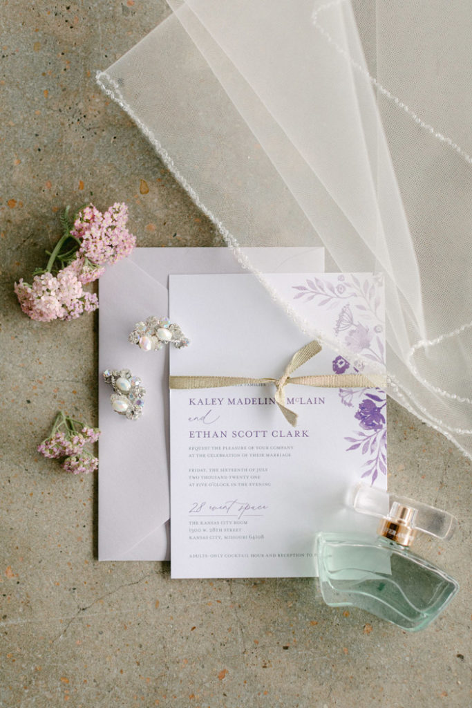 QCB Wedding invitation with purple flowers and lavender envelope, styled with earrings, veil, and perfume