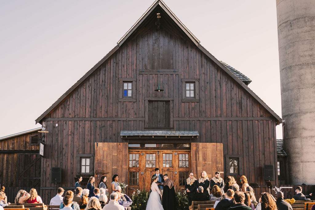 Bride and Groom stand in front of large barn doors with woodsy floral installation at their feet - Horizontal