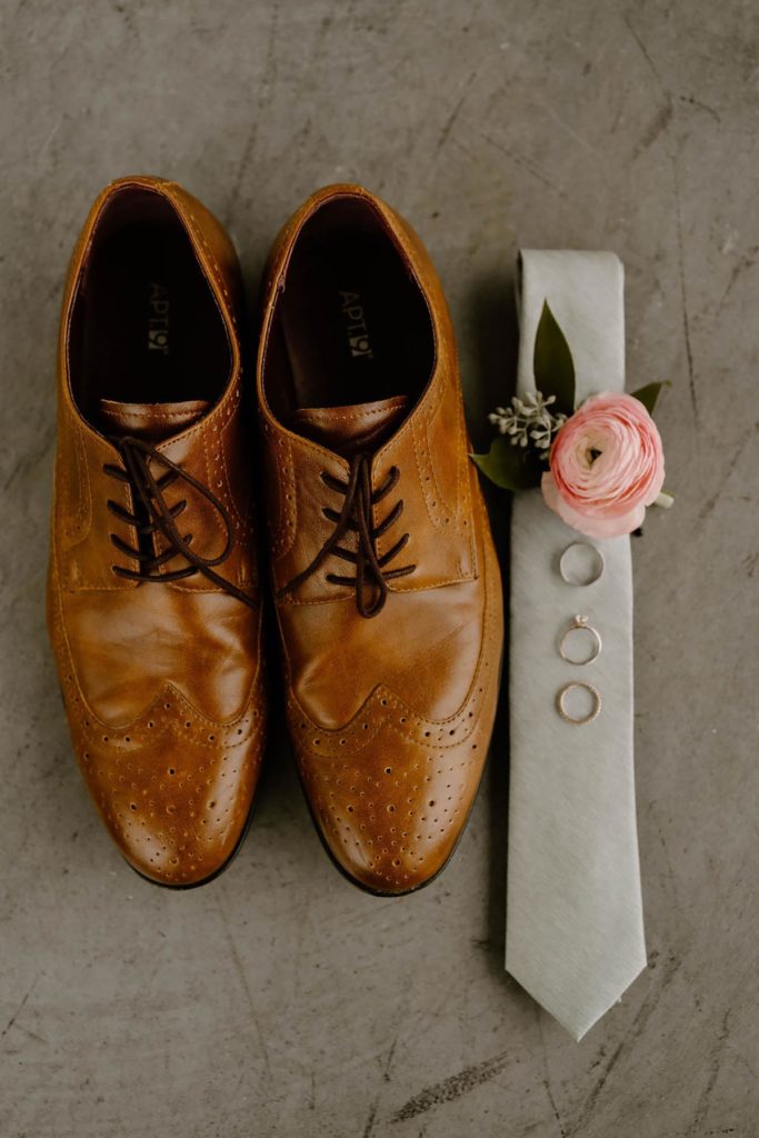 Peach boutonniere laying on a gray tie with wedding rings, and leather shoes sitting to the left