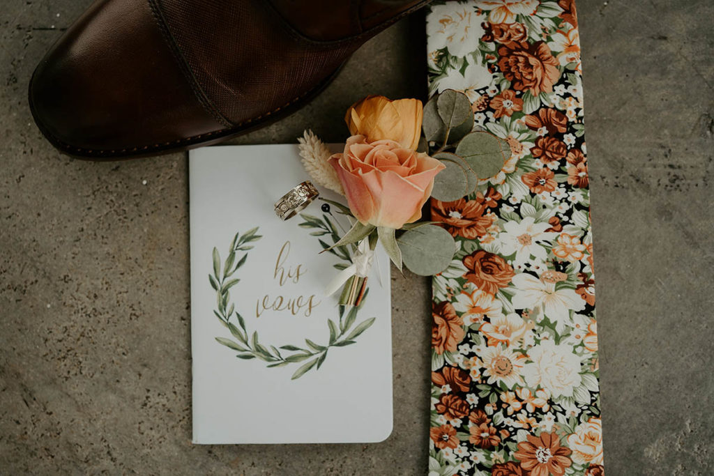 Flat lay of the groom's vow book, shoes, tie, and boutonniere
