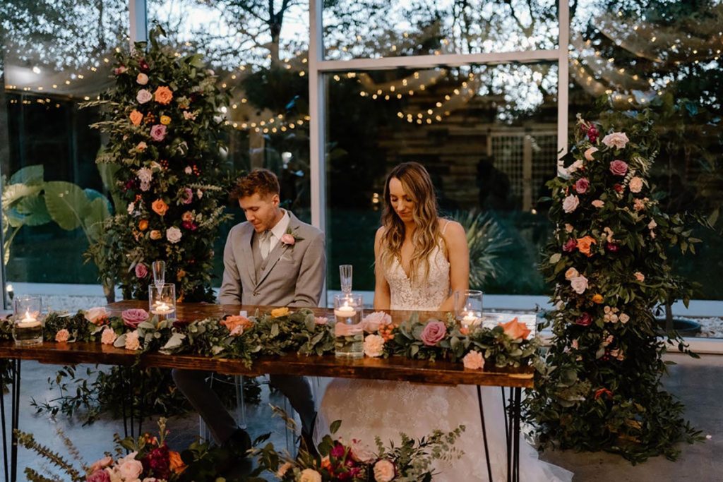 Couple sitting at the sweetheart table with floral arrangements in front of the table, on the table, and floral pillars behind the table