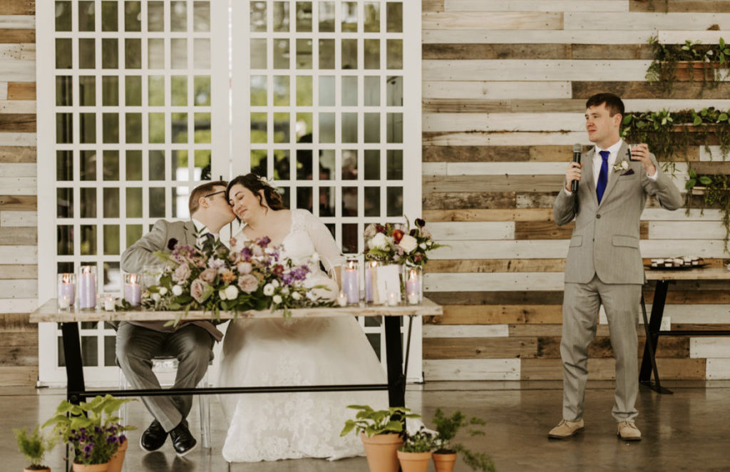 Groom kissing bride's cheek while sitting at the sweetheart table