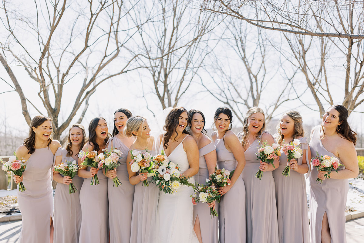 Bride in the center of her bridesmaids laughing and holding muted pastel bouquets