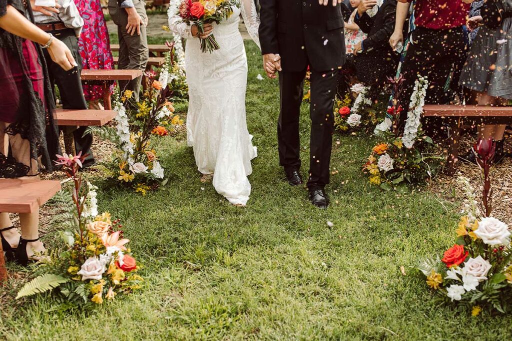 Bride and groom walk past aisle florals while guests toss petals at them like confetti