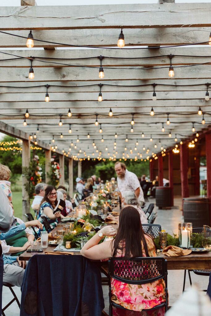 Large outdoor banquet table with guests seated around it and bistro lights hanging above.