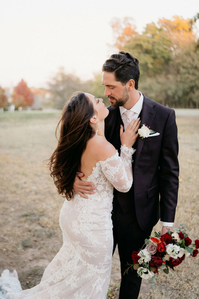 Groom holds bouquet as they gaze at each other with fall background