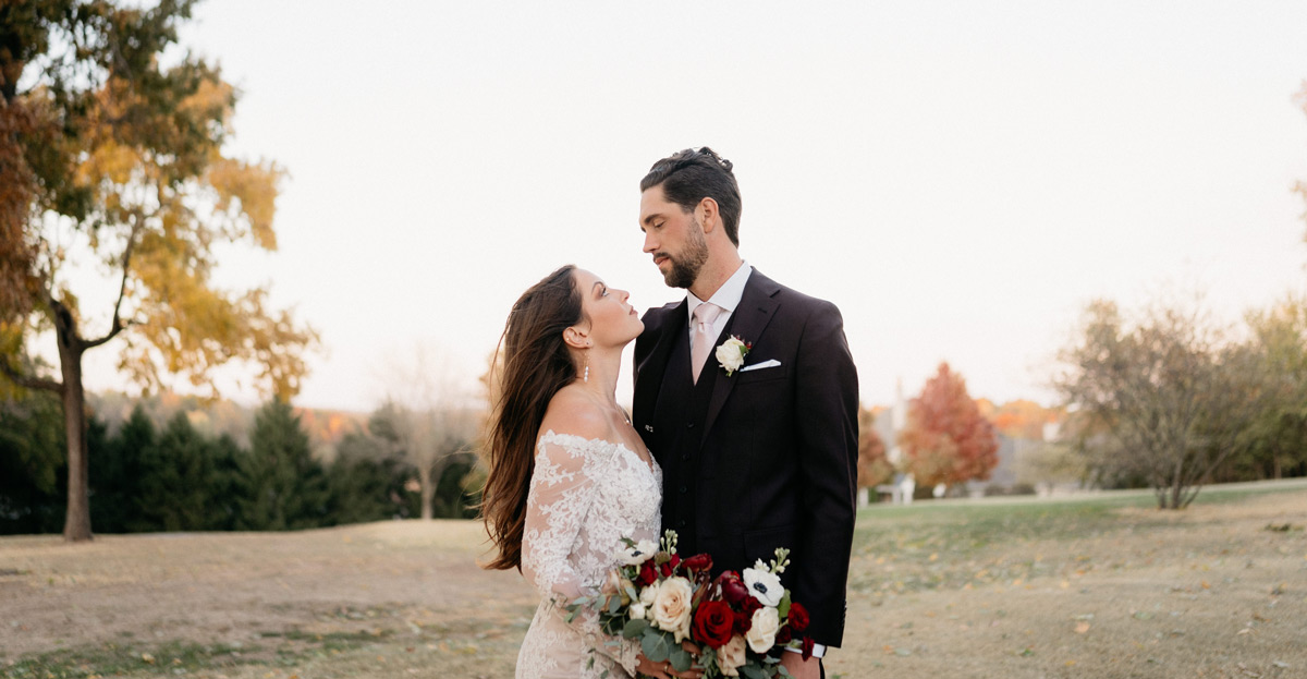 Bride and Groom gazing at each other in front of fall tree line, holding burgundy bridal bouquet
