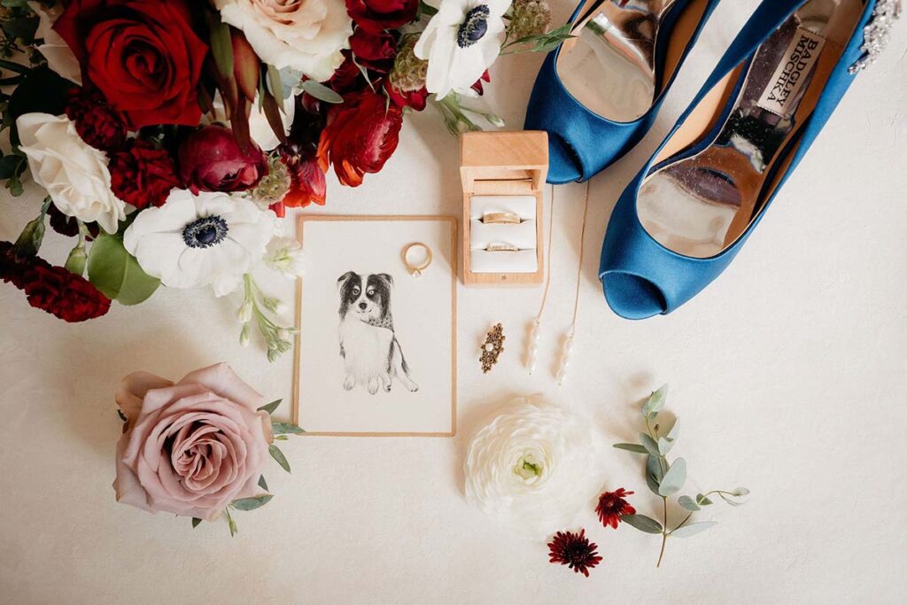 Flat lay with bouquet, brides' shoes, rings, jewelry, and illustration of their pup to be used as their sweetheart table marker.