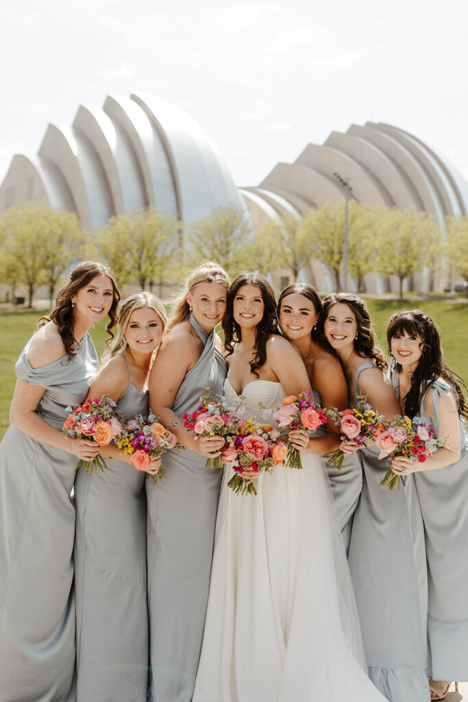 Bride in center with bridesmaids on either side smiling, all holding bouquets with the Kauffman Center in the background