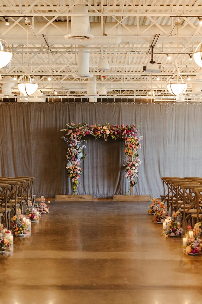 Full floral rectangle arbor at the front of the room with candles and colorful arrangements at the base of the chairs in the aisle