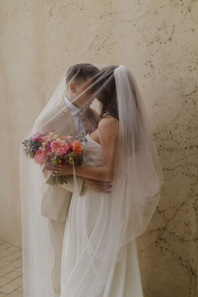 Bride and groom holding bouquet with veil draped romantically over them.