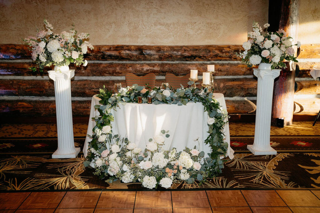 Sweetheart table with a cascading garland, and flowers at the foot of the table, as well as on columns on either side of the table