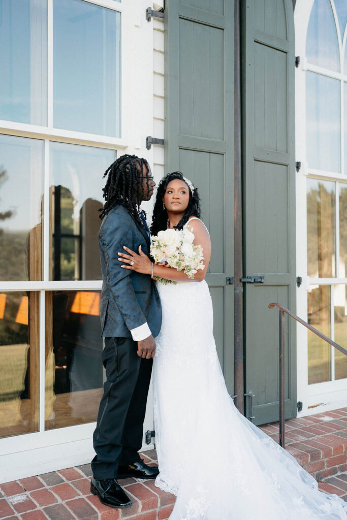 Groom and bride posing together in front of large chapel windows, looking into distance